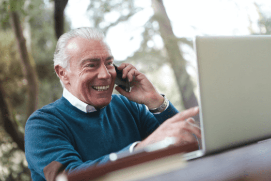A smiling, elderly man talking on the phone and pointing at his laptop.