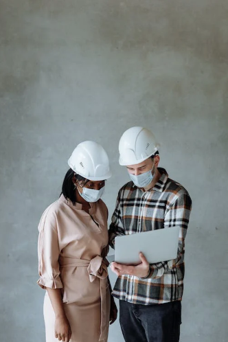 A black woman and Caucasian man wearing hard hats looking at an open laptop