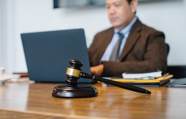 A gavel on a wooden desk in front of a solicitor typing on a laptop.